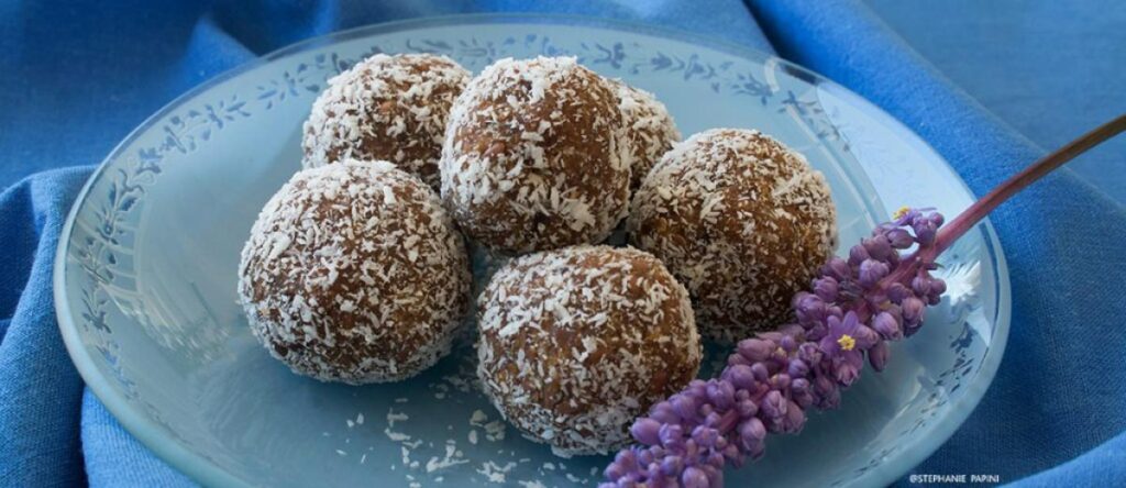 Coconut-coated date energy balls on a light blue glass plate next to a purple flower branch.
