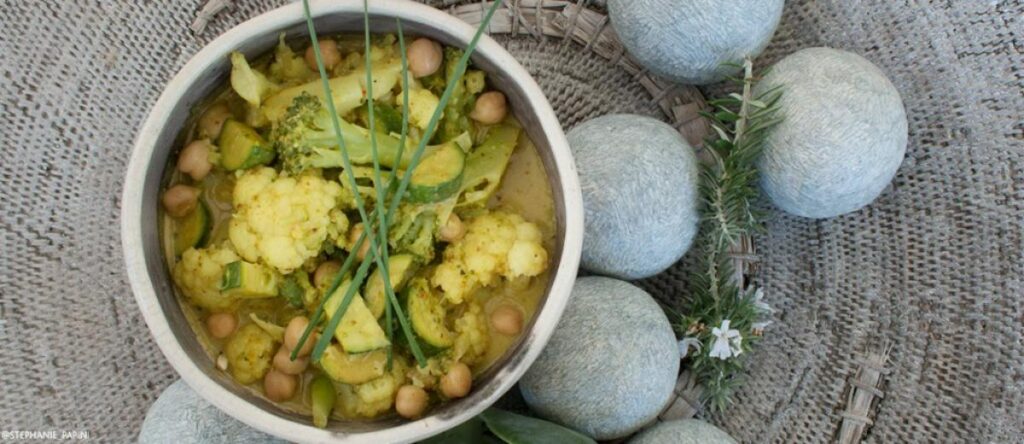 Coconut cauliflower curry with broccoli, chickpeas, and zucchini in a white bowl on a woven placemat.