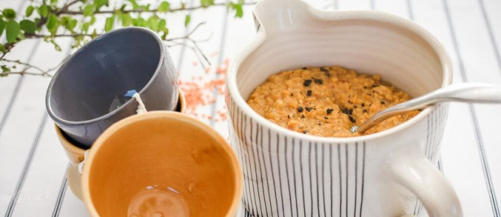 Creamy orange lentil dhal with black spice garnishes in a white striped ceramic jug next to colorful bowls.