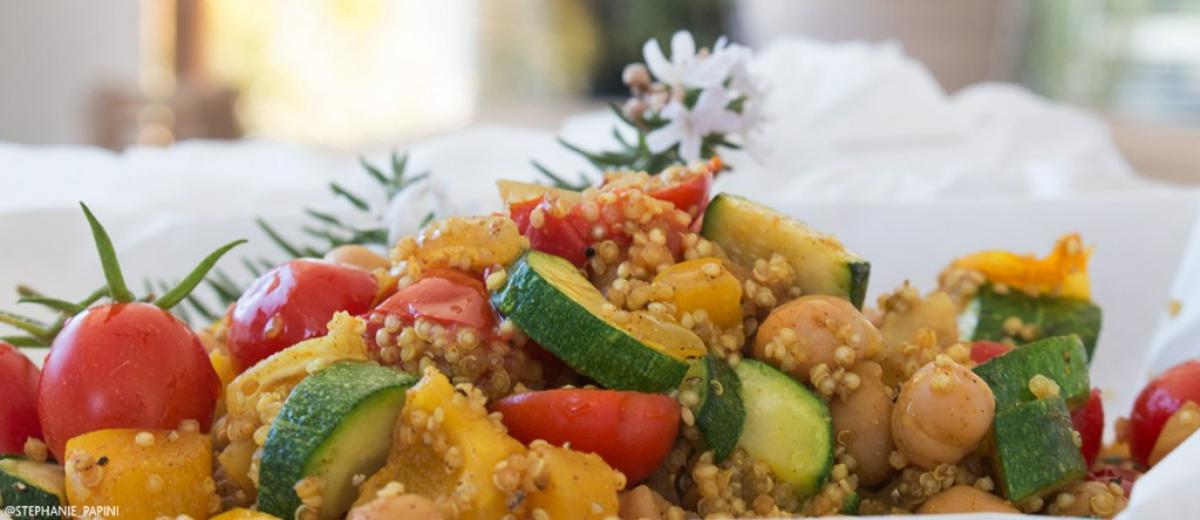 A bowl of curried quinoa with halved cherry tomatoes, sliced zucchini, and chickpeas on a white background.