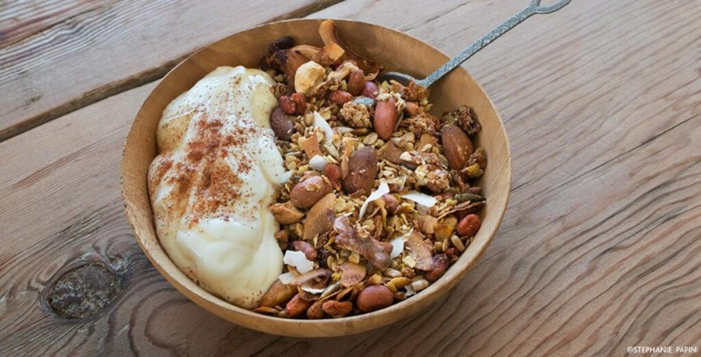 A wooden bowl of homemade granola topped with yogurt and a dusting of cinnamon on a rustic wooden table.