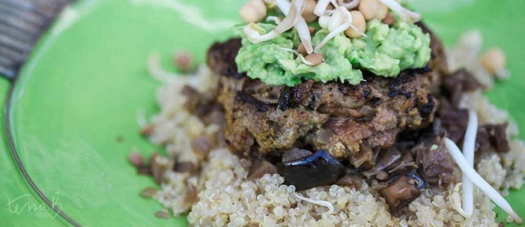 A Moroccan beef burger patty topped with avocado and sprouts on a bed of quinoa and aubergine in a green bowl.