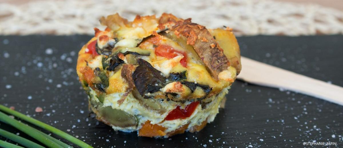 A muffin-sized frittata with melted cheese and visible red peppers on a black slate board with chives.