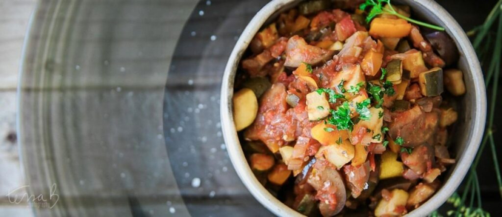 A close-up of a colorful vegetable ratatouille with diced aubergine, tomatoes, and herbs in a white ceramic bowl.