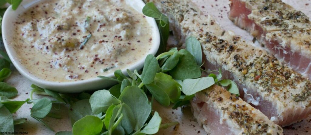Sliced seared tuna steaks with green herbs next to a small bowl of creamy pistachio dressing on a textured plate.