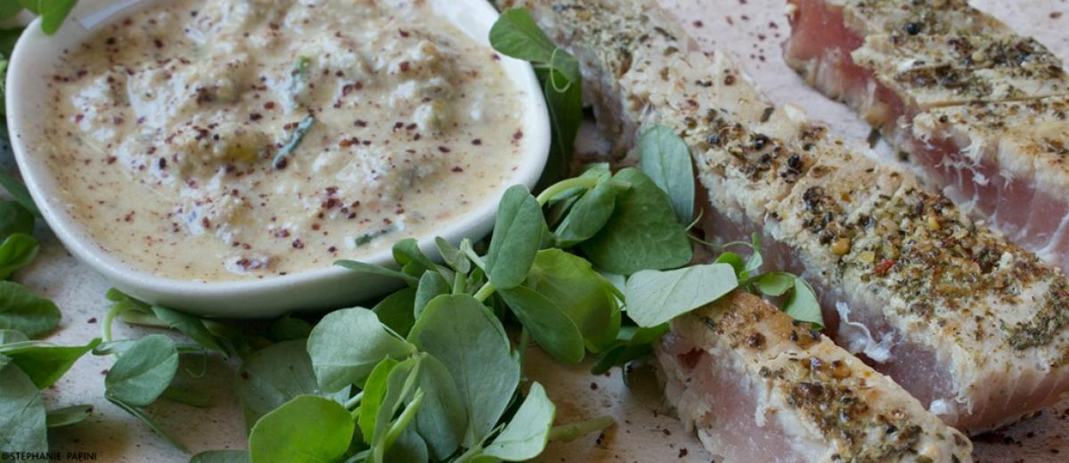 Sliced seared tuna steaks with green herbs next to a small bowl of creamy pistachio dressing on a textured plate.