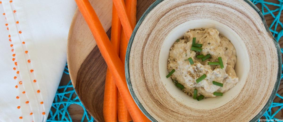A ceramic bowl of hummus topped with fresh chives, served with long orange carrot sticks on a wooden board