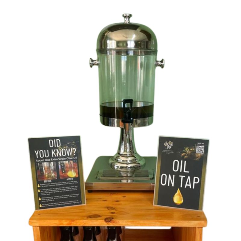 A stainless steel and glass extra virgin olive oil on tap dispenser unit with informational display signs.